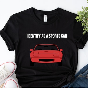 Op de afbeelding: Zwart t-shirt met de tekst "I IDENTIFY AS A SPORTS CAR" boven een rode autografiek. De autografiek is een vooraanzicht van een sportwagen. Het shirt is gemaakt van een zacht materiaal.