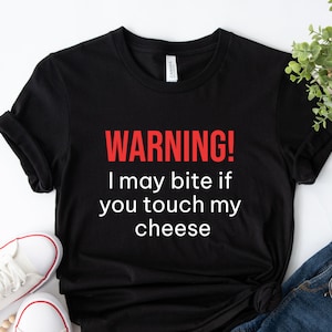 以下が含まれることがあります： 「WARNING! I may bite if you touch my cheese」のテキストが赤と白でプリントされた黒いTシャツ。白いスニーカーとデニムジーンズと一緒に表示されています。