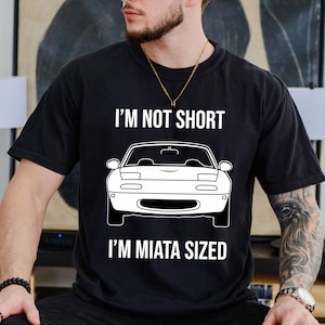May include: Black t-shirt with the text "I'M NOT SHORT I'M MIATA SIZED" above and below a white car graphic. The shirt is a crew neck and appears to be made of cotton.
