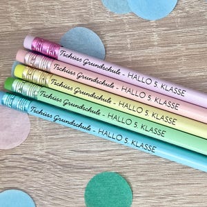 Könnte beinhalten: Ein Set aus vier pastellfarbenen Bleistiften in Rosa, Gelb, Hellgrün und Hellblau, jeweils mit einer Metallzwinge mit Radiergummi und dem aufgedruckten schwarzen Text Tschüss Grundschule - HALLO 5. KLASSE, die diagonal gestapelt sind.