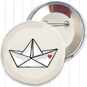 Könnte beinhalten: Ein runder Ansteckbutton mit einer Illustration eines weißen Papierboots, das an der Seite mit einem kleinen roten Herz versehen ist. Der Button hat einen cremefarbenen Hintergrund und eine silberne Metallrückseite mit einer Nadel.