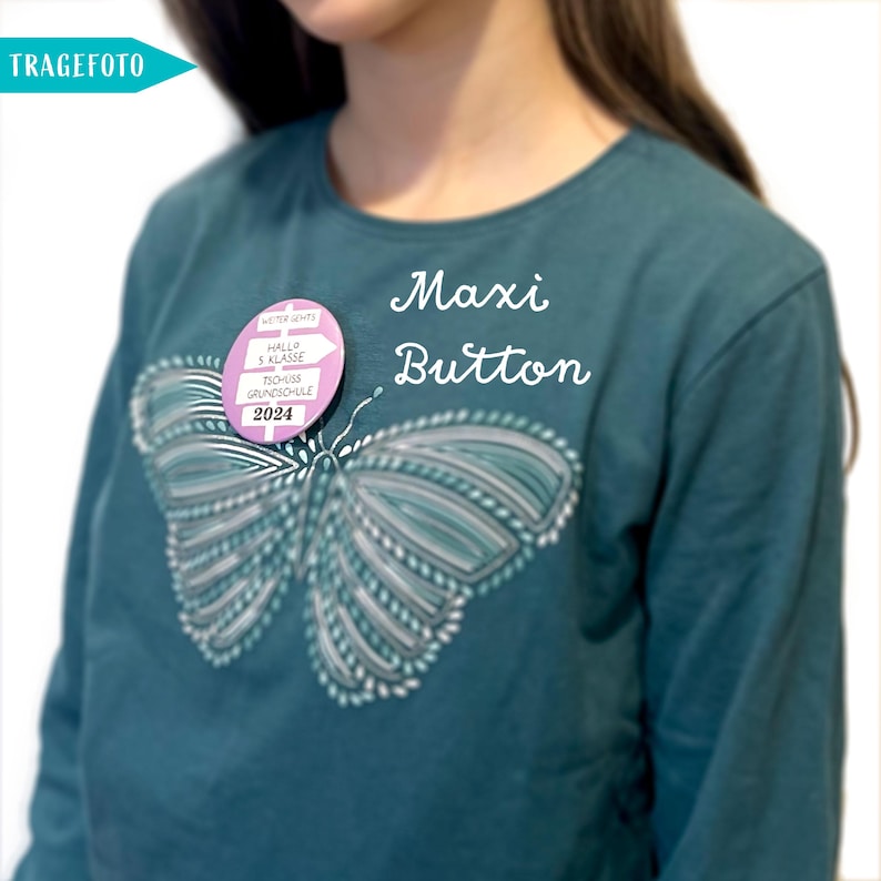 May include: A teal shirt with a butterfly design and a pink button that says "Weiter geht's" with an arrow pointing right, "Hallo 5 Klasse", "Tisch 15", "Grundschule", and "2024". The text "Maxi Button" is written in cursive on the shirt.