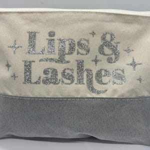 Könnte beinhalten: Eine cremefarbene Kosmetiktasche mit grauem Boden und Reißverschluss. Die Worte "Lips & Lashes" sind in silbernem Glitzer auf der Vorderseite mit Sternakzenten geschrieben.