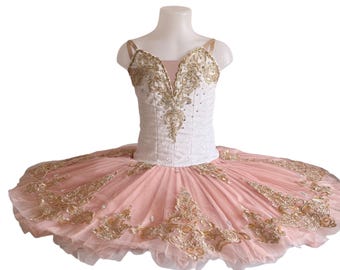 May include: A ballet tutu with a white bodice and a pink skirt. The bodice is embellished with gold embroidery and rhinestones. The skirt is layered and has gold embroidery along the edges. The straps are a light tan color.