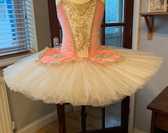May include: A pink and white ballet tutu with gold embroidery and embellishments. The tutu is on a stand and has a full skirt.