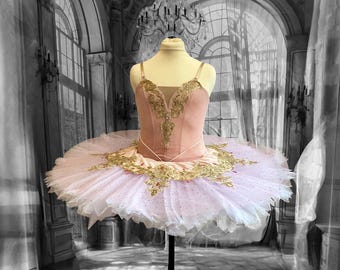 May include: A pink and white tulle ballet tutu with gold embroidery and embellishments. The tutu is on a dress form in a room with a chandelier.