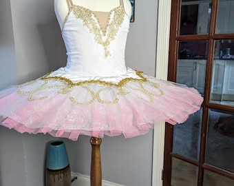May include: A white and pink ballet tutu with gold trim and embellishments. The tutu is on a wooden stand.