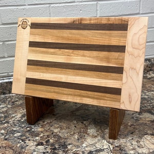 May include: A wooden cutting board with a stand, made of maple and walnut wood. The board has a natural wood finish and is perfect for serving cheese, charcuterie, or other appetizers.