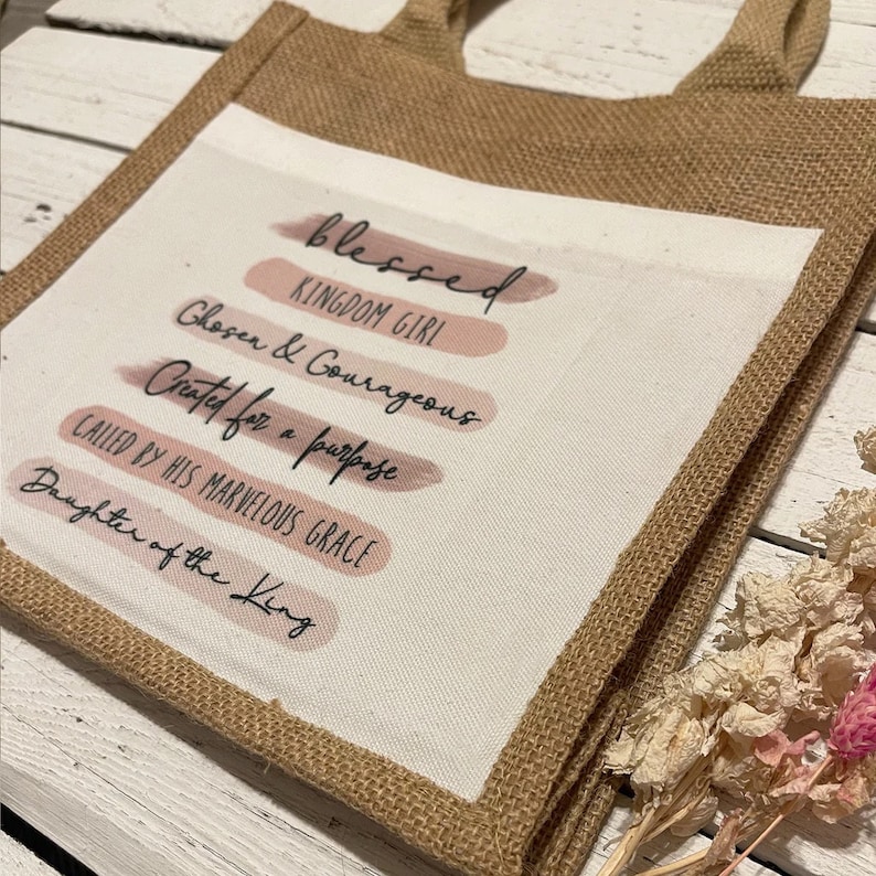 Pode incluir: Uma bolsa de juta com um inserto de lona branca que apresenta o texto "blessed", "kingdom girl", "chosen & courageous", "created for a purpose", "called by his marvelous grace" e "daughter of the king" em letras pretas. O texto &eacute; sobreposto a um fundo de aquarela rosa.