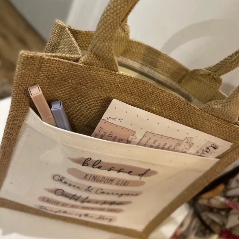 Pode incluir: Uma bolsa de juta marrom com um bolso branco que apresenta o texto "blessed kingdom girl" e as frases "Chosen & Courageous", "Created for a purpose", "Called by His name" e "Daughter of the King".