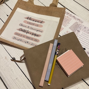 Pode incluir: Uma bolsa de juta com um inserto de lona branca que apresenta o texto "blessed kingdom girl chosen & courageous created for a purpose called by his marvelous grace daughter of the king". O texto est&aacute; em rosa e preto. A bolsa est&aacute; sobre uma superf&iacute;cie de madeira com um di&aacute;rio de couro marrom, tr&ecirc;s canetas e um bloco de notas rosa.