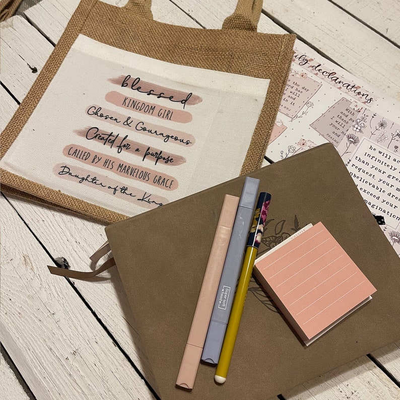 Pode incluir: Uma bolsa de juta com um inserto de lona branca que apresenta o texto "blessed kingdom girl chosen & courageous created for a purpose called by his marvelous grace daughter of the king". O texto est&aacute; em rosa e preto. A bolsa est&aacute; sobre uma superf&iacute;cie de madeira com um di&aacute;rio de couro marrom, tr&ecirc;s canetas e um bloco de notas rosa.