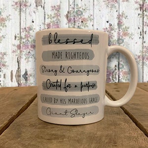May include: White ceramic mug with a grey and black design. The mug has the words "blessed", "made righteous", "strong & courageous", "created for a purpose", "called by his marvellous grace", and "giant slayer" printed on it.