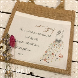 Puede incluir: Una bolsa de mano de yute con un inserto de lona blanca que presenta un diseño floral y la cita "Ella está vestida de fuerza y dignidad, se ríe sin miedo al futuro. Prov 31:25".