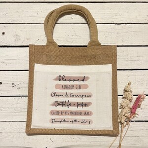 Pode incluir: Uma bolsa de juta marrom com um inserto de lona branca. O inserto de lona tem o texto "blessed" em preto com listras de aquarela rosa por baixo. O texto "Kingdom Girl" est&aacute; em preto abaixo da palavra "blessed". O texto "Chosen & Courageous" est&aacute; em preto abaixo de "Kingdom Girl". O texto "Created for a purpose" est&aacute; em preto abaixo de "Chosen & Courageous". O texto "Called by His Marvelous Grace" est&aacute; em preto abaixo de "Created for a purpose". O texto "Daughter of the King" est&aacute; em preto abaixo de "Called by His Marvelous Grace".