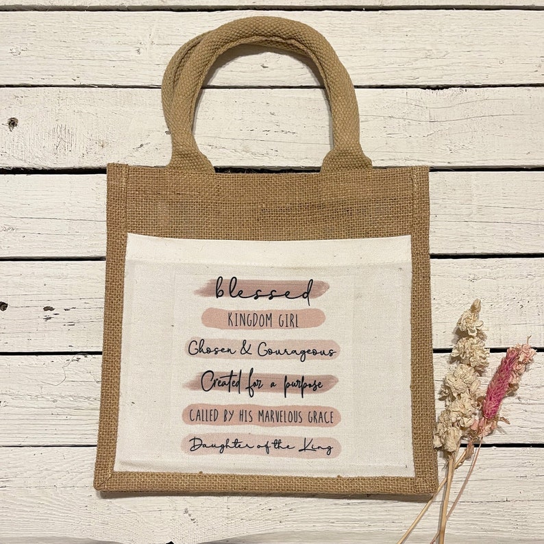 Pode incluir: Uma bolsa de juta marrom com um inserto de lona branca. O inserto de lona tem o texto "blessed" em preto com listras de aquarela rosa por baixo. O texto "Kingdom Girl" est&aacute; em preto abaixo da palavra "blessed". O texto "Chosen & Courageous" est&aacute; em preto abaixo de "Kingdom Girl". O texto "Created for a purpose" est&aacute; em preto abaixo de "Chosen & Courageous". O texto "Called by His Marvelous Grace" est&aacute; em preto abaixo de "Created for a purpose". O texto "Daughter of the King" est&aacute; em preto abaixo de "Called by His Marvelous Grace".