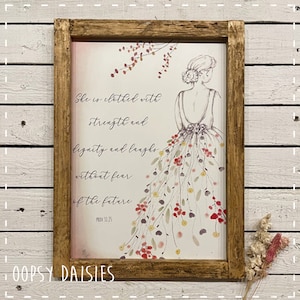 May include: A framed print with a watercolor illustration of a woman in a floral dress. The text reads "She is clothed with strength and dignity and laughs without fear of the future. Prov 31:25".
