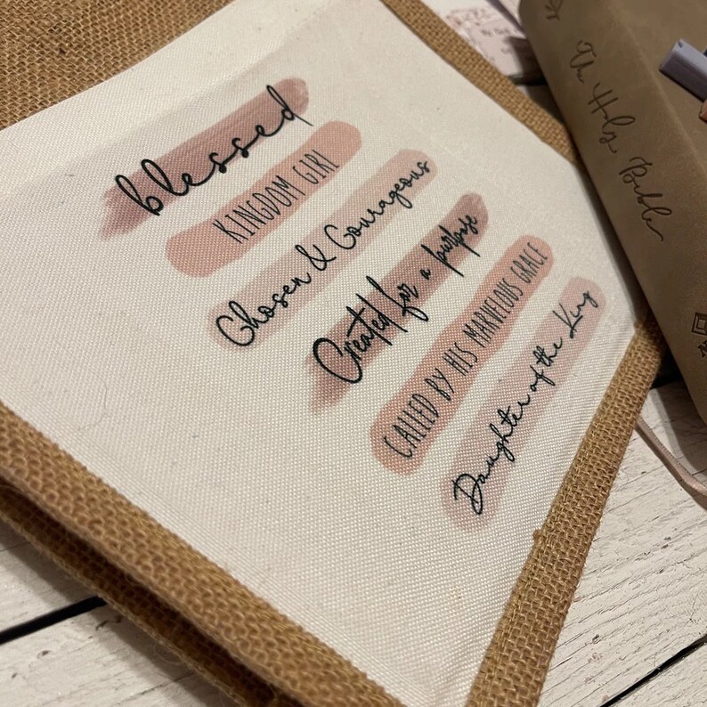 Pode incluir: Uma bolsa tote de lona bege com uma borda de juta. A bolsa tem listras de aquarela rosa com o texto "blessed", "kingdom girl", "chosen & courageous", "created for a purpose", "called by his marvelous grace" e "daughter of the king" em preto.