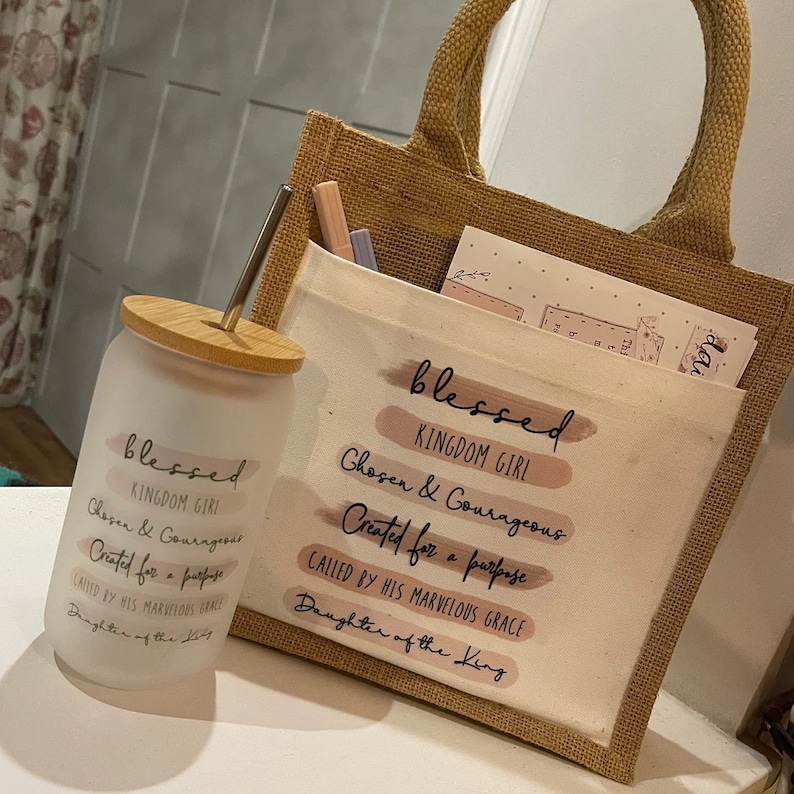 Pode incluir: Uma bolsa tote de juta com um inserto de lona branca que apresenta o texto "blessed kingdom girl chosen & courageous created for a purpose called by his marvelous grace daughter of the king". A bolsa &eacute; combinada com um copo de vidro transparente com tampa de madeira e um canudo. O copo tem o mesmo texto que a bolsa.
