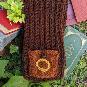 Può includere: Una borsa di maglia marrone con una piccola tasca sul davanti. La tasca ha un bottone marrone e un anello dorato ricamato su di essa.