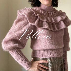 Puede incluir: Un suéter de color rosa empolvado con cuello alto y cuello con volantes. El suéter tiene mangas largas con puños acanalados y una cintura acanalada. La palabra "Pattern" está escrita en la parte delantera del suéter.