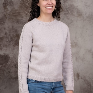Peut inclure: Un pull en tricot à manches longues de couleur lavande clair, doté d'une encolure ronde et de détails en torsades verticales le long de la partie extérieure de chaque manche. Le pull présente un ourlet et des poignets côtelés, porté avec un jean bleu.