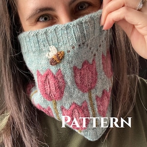 Pode incluir: Um cachecol de malha azul claro com um padrão de tulipas cor-de-rosa e uma abelha. O cachecol está sendo segurado pela mão de uma pessoa, e a palavra "PATTERN" é visível na parte inferior da imagem.