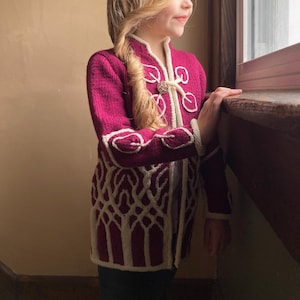 May include: A magenta knitted jacket with white trim and a decorative clasp. The jacket features a unique white pattern on the lower portion and around the cuffs. The long-sleeved jacket is a stylish outerwear option.
