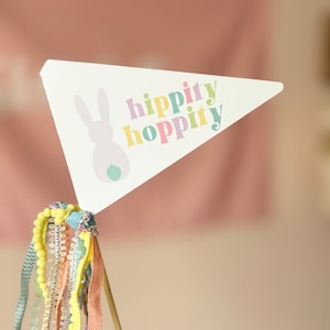 May include: A white triangular flag with a bunny silhouette and the text "hippity hoppity" in rainbow colours. The flag is attached to a wooden stick with colourful ribbons.