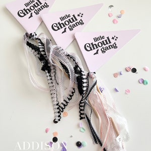 May include: Three light pink pennant flags with the text "little Ghoul gang" in black. The flags are decorated with black bats and stars. The flags are attached to a bundle of ribbons in black, white, and pink. Confetti is scattered around the flags.