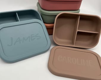 May include: Four silicone lunch boxes with lids. The boxes are in different colors: pink, green, brown, and blue. The lids have names on them: "JAMES" and "CAROLINE".