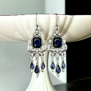 May include: Silver-toned chandelier earrings with a square, dark blue gemstone center and smaller teardrop-shaped blue gems. The earrings have a decorative filigree design and are displayed on a white pedestal.