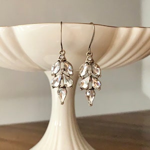 May include: A pair of silver-toned drop earrings with clear, faceted crystal accents. The earrings feature a leaf-like design with multiple crystal shapes, including teardrops. The earrings are displayed on a white pedestal cake stand.
