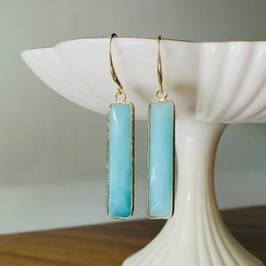 May include: A pair of gold-toned earrings with rectangular, light blue pendants. The earrings are displayed on a white, tiered pedestal. The pendants have a gold border and a smooth, polished appearance. The earrings are designed with a hook closure.