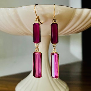 May include: A pair of gold-toned earrings with rectangular, magenta-colored gems. The earrings are displayed on a white, tiered pedestal. The gems have a gradient effect, transitioning from a darker magenta to a lighter pink.