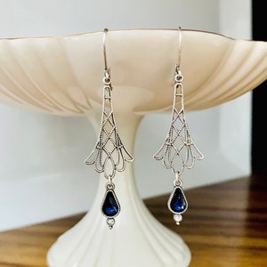 May include: A pair of silver-toned drop earrings. Each earring features an openwork design with a teardrop-shaped, dark blue gemstone and a small, clear accent stone. The earrings are displayed on a white, tiered pedestal.