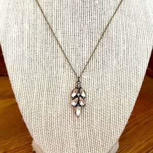 May include: A delicate necklace with a pendant featuring clear, teardrop-shaped crystals. The pendant is suspended from a thin, antique-bronze colored chain. The necklace is displayed on a cream-colored fabric.