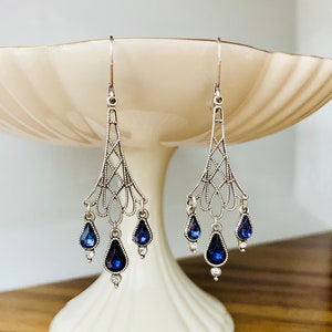 May include: Silver-toned chandelier earrings with a filigree design. Each earring features a series of dangling, teardrop-shaped, deep blue gemstones and small clear crystals. The earrings are displayed on a white pedestal.