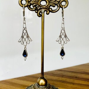 May include: Silver-toned dangle earrings with a filigree design, featuring a teardrop-shaped, dark blue gemstone. The earrings are displayed on a decorative, antique-style stand with a brass finish.