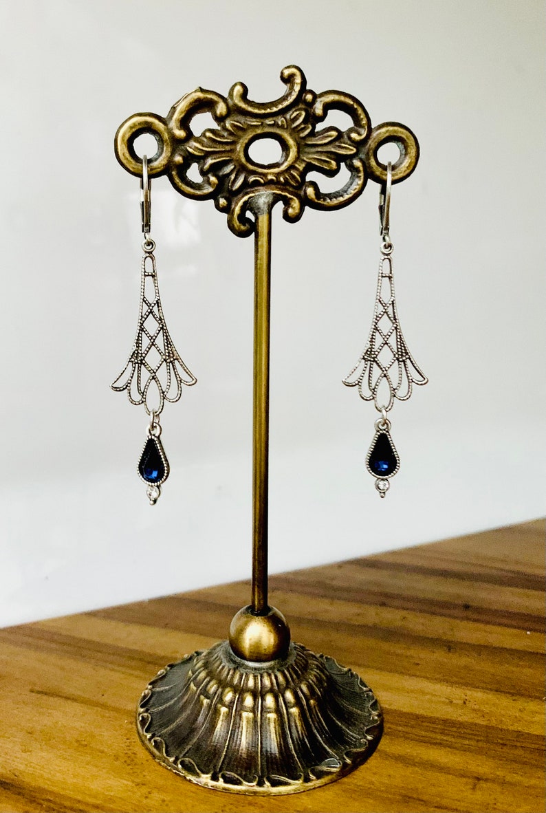May include: Silver-toned dangle earrings with a filigree design, featuring a teardrop-shaped, dark blue gemstone. The earrings are displayed on a decorative, antique-style stand with a brass finish.