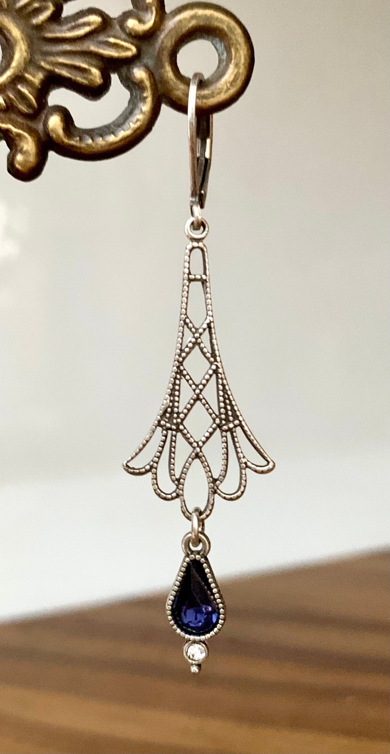 May include: Silver-toned filigree earring with a teardrop-shaped, dark blue gemstone and a small, round, clear stone. The earring has a decorative, openwork design with a lever-back closure. The earring is suspended from a decorative metal fixture.