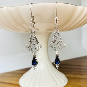 May include: A pair of silver-toned drop earrings. Each earring features an openwork design with a teardrop-shaped, dark blue gemstone and a small, clear accent stone. The earrings are displayed on a white, tiered pedestal.