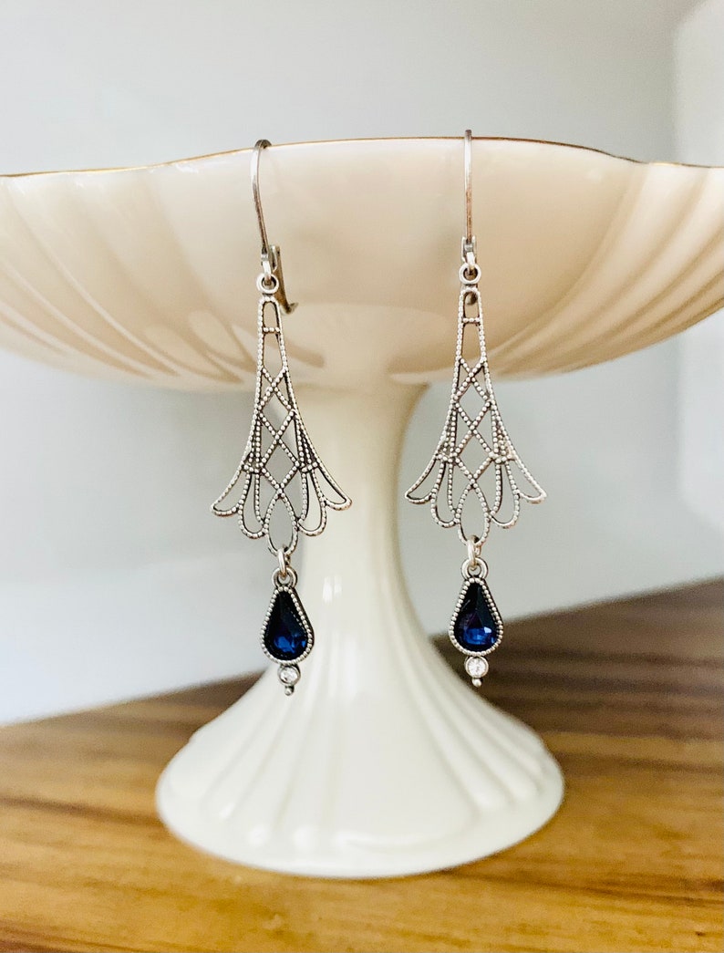 May include: A pair of silver-toned drop earrings. Each earring features an openwork design with a teardrop-shaped, dark blue gemstone and a small, clear accent stone. The earrings are displayed on a white, tiered pedestal.