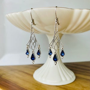 May include: Silver-toned chandelier earrings with a delicate filigree design. Each earring features three teardrop-shaped, deep blue gemstones. The earrings are displayed on a white pedestal dish against a light background.
