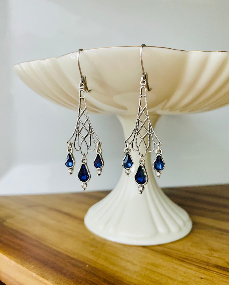May include: Silver-toned chandelier earrings with a delicate filigree design. Each earring features three teardrop-shaped, deep blue gemstones. The earrings are displayed on a white pedestal dish against a light background.