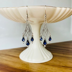 May include: Silver-toned chandelier earrings with intricate filigree design. Each earring features three teardrop-shaped, deep blue gemstones and small clear accents. The earrings are displayed on a white pedestal.