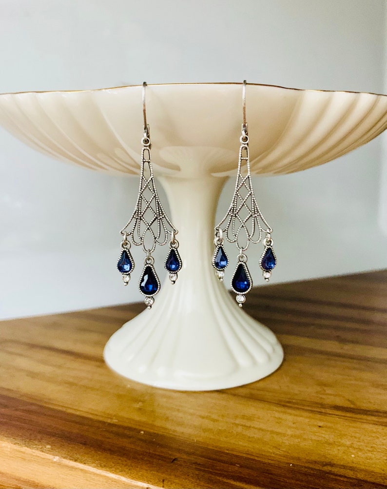May include: Silver-toned chandelier earrings with intricate filigree design. Each earring features three teardrop-shaped, deep blue gemstones and small clear accents. The earrings are displayed on a white pedestal.