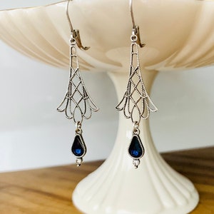 May include: Silver-toned drop earrings with an Art Deco design. Each earring features a filigree top with a geometric pattern and a teardrop-shaped, dark blue gemstone pendant. The earrings are displayed on a white pedestal.