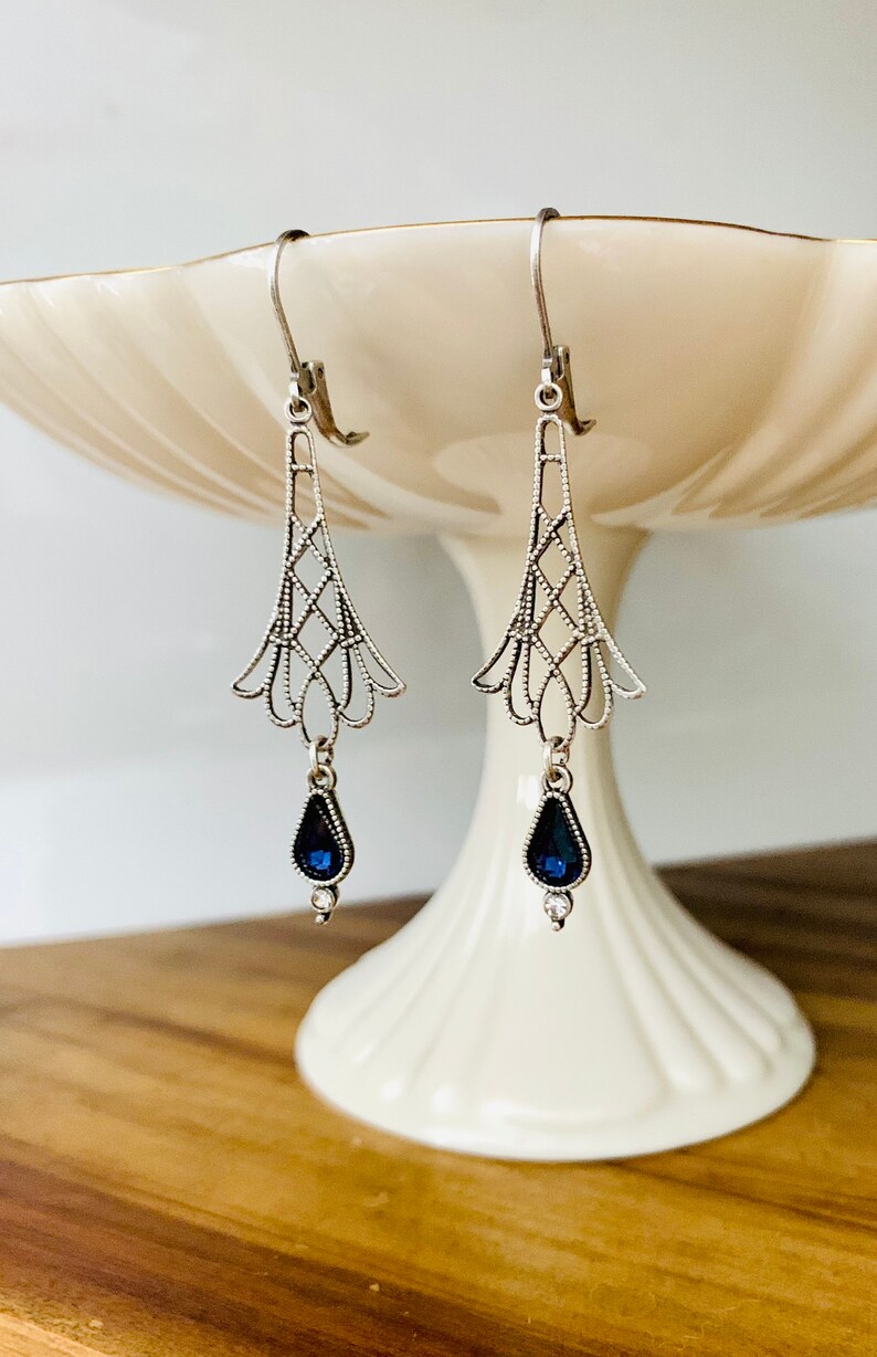 May include: Silver-toned drop earrings with an Art Deco design. Each earring features a filigree top with a geometric pattern and a teardrop-shaped, dark blue gemstone pendant. The earrings are displayed on a white pedestal.