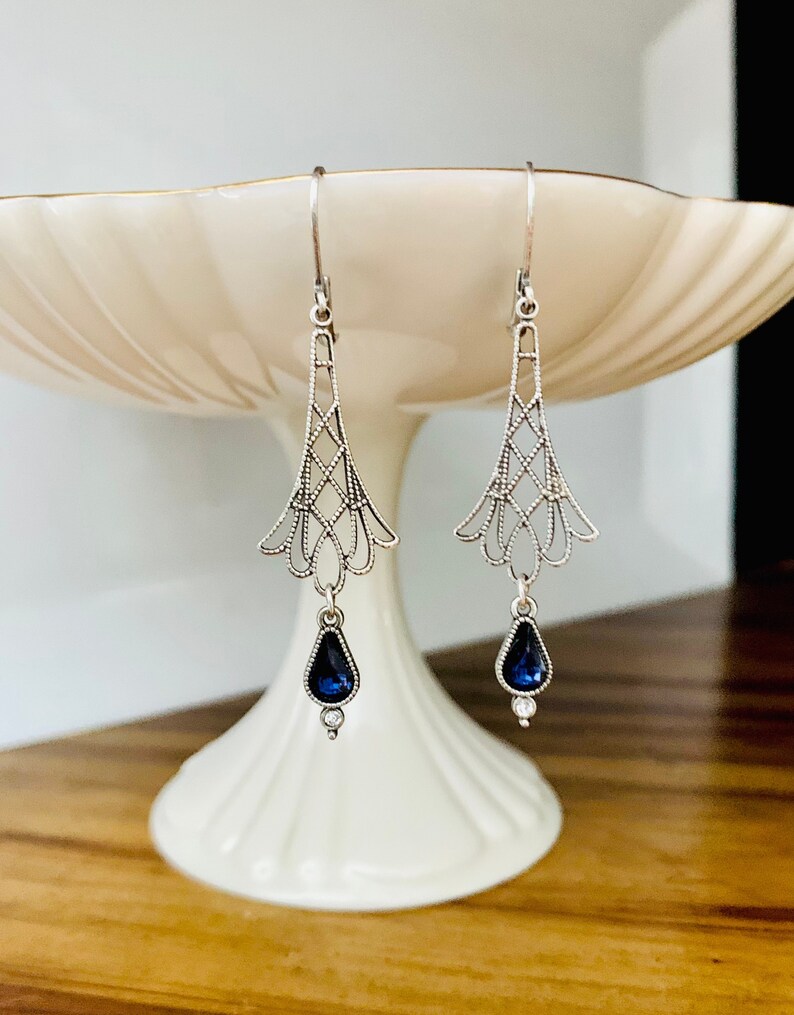 May include: A pair of silver-toned drop earrings. Each earring features an openwork design with a teardrop-shaped, dark blue gemstone and a small, clear accent stone. The earrings are displayed on a white, tiered pedestal.
